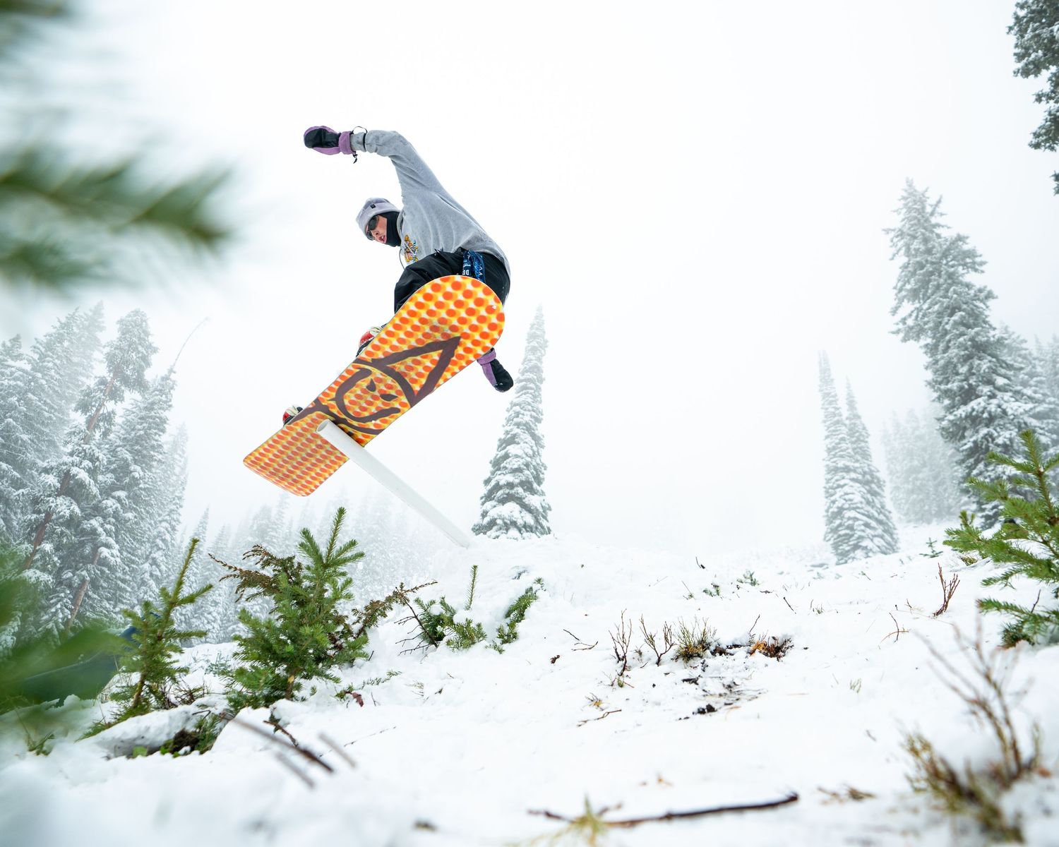 Snowboarder performing a dramatic aerial trick with snow-covered evergreens in the background.