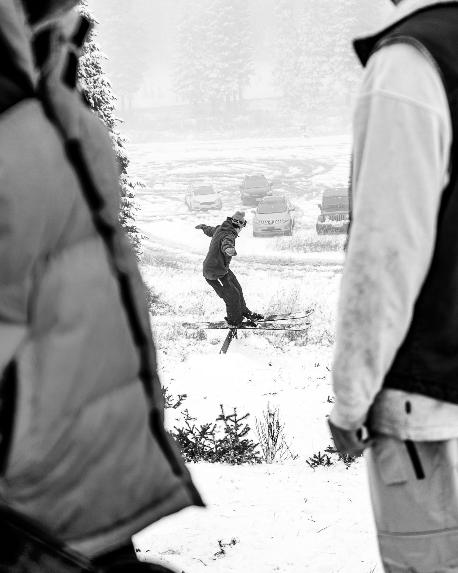 Black and white image of a skier jumping through winter mountain terrain with observers in foreground.