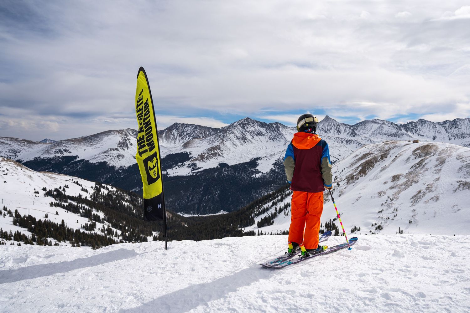 Skier in orange jacket stands at mountain peak with yellow banner against snowy landscape.