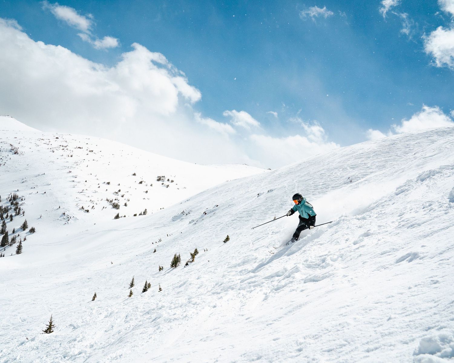 Lone skier descending a vast snow-covered mountain slope under blue skies.