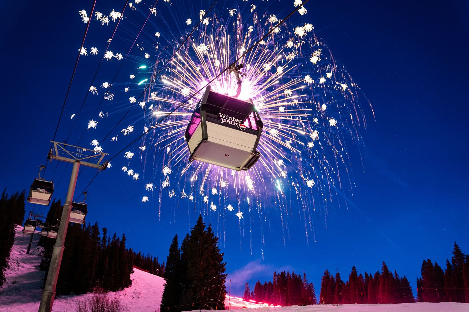 Fireworks explode behind a ski lift gondola against dark blue winter sky.