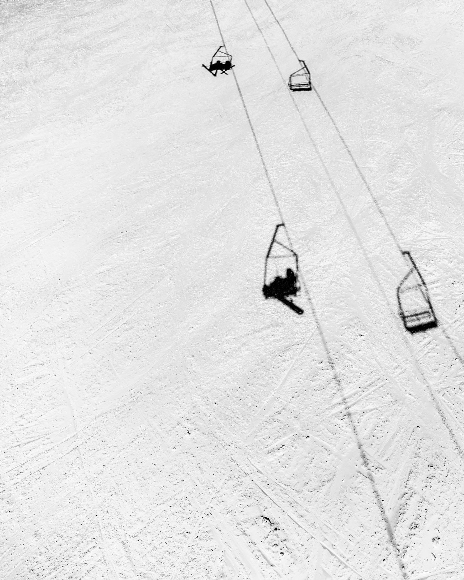 Aerial view of ski lift chairs casting shadows on snowy slope in black and white.