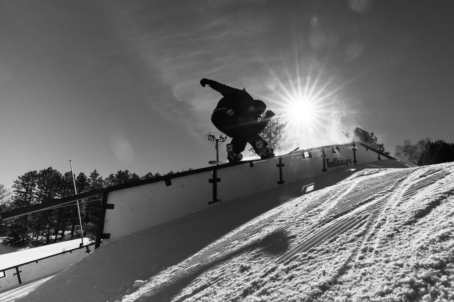 Snowboarder performs a rail grind trick on a snowy slope with bright sun flare in black and white photography.