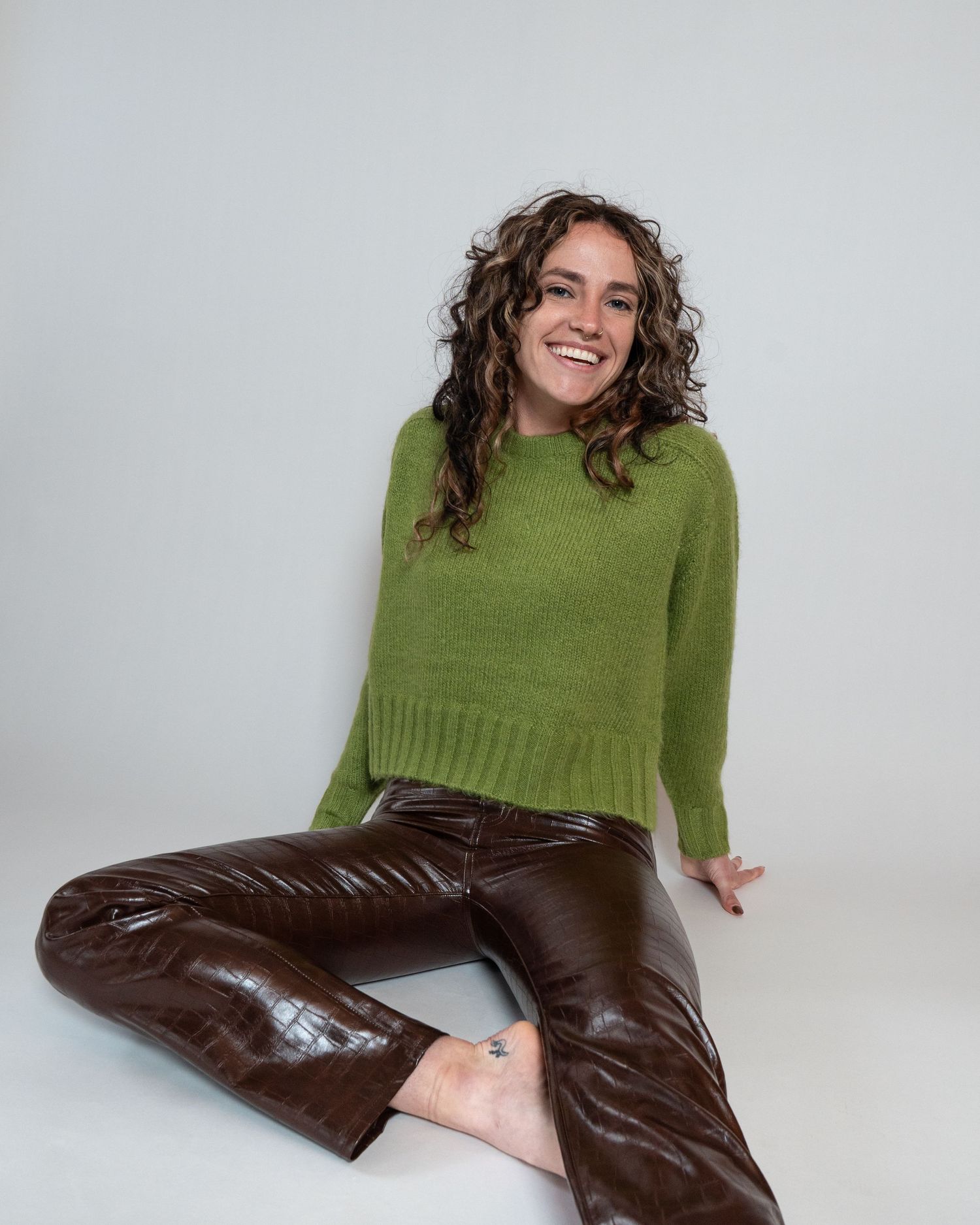 Person wearing a green knit sweater and brown leather pants sits casually on white background.