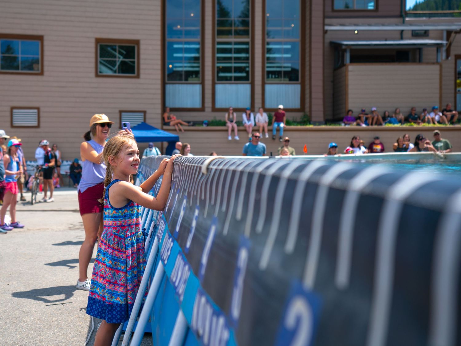 Spectators gather around a large blue barricaded pool area during an outdoor summer event.