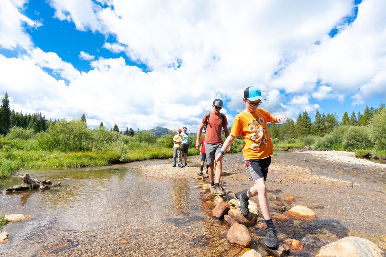 Hikers crossing a stream using stepping stones in a mountain landscape with evergreen trees.