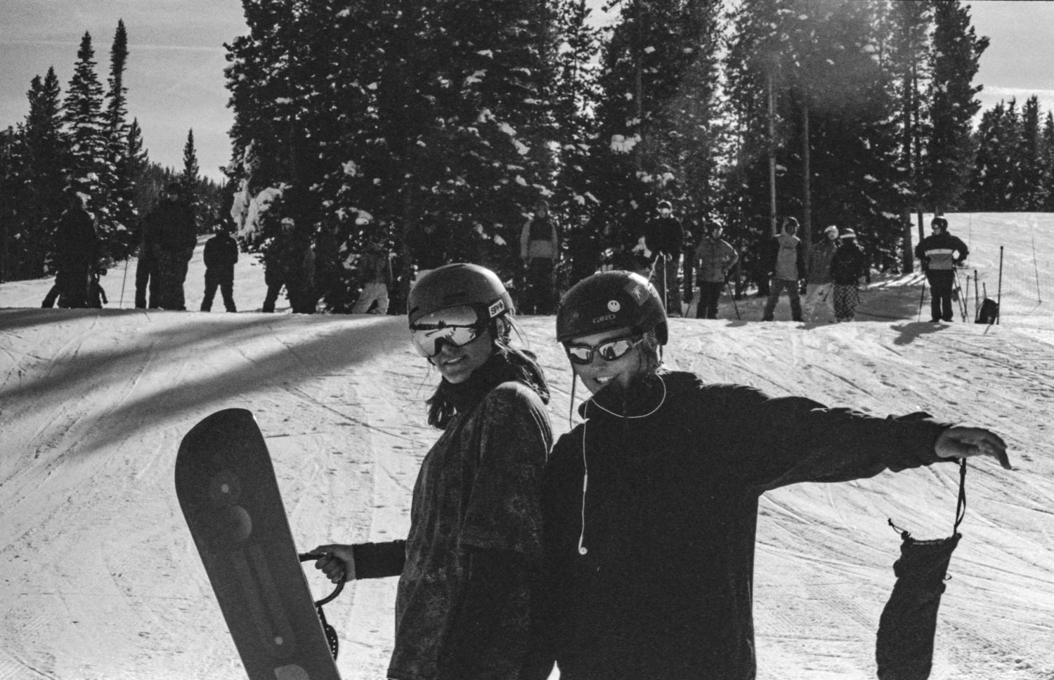 Two snowboarders pose together on snowy slopes with evergreen trees in the background.
