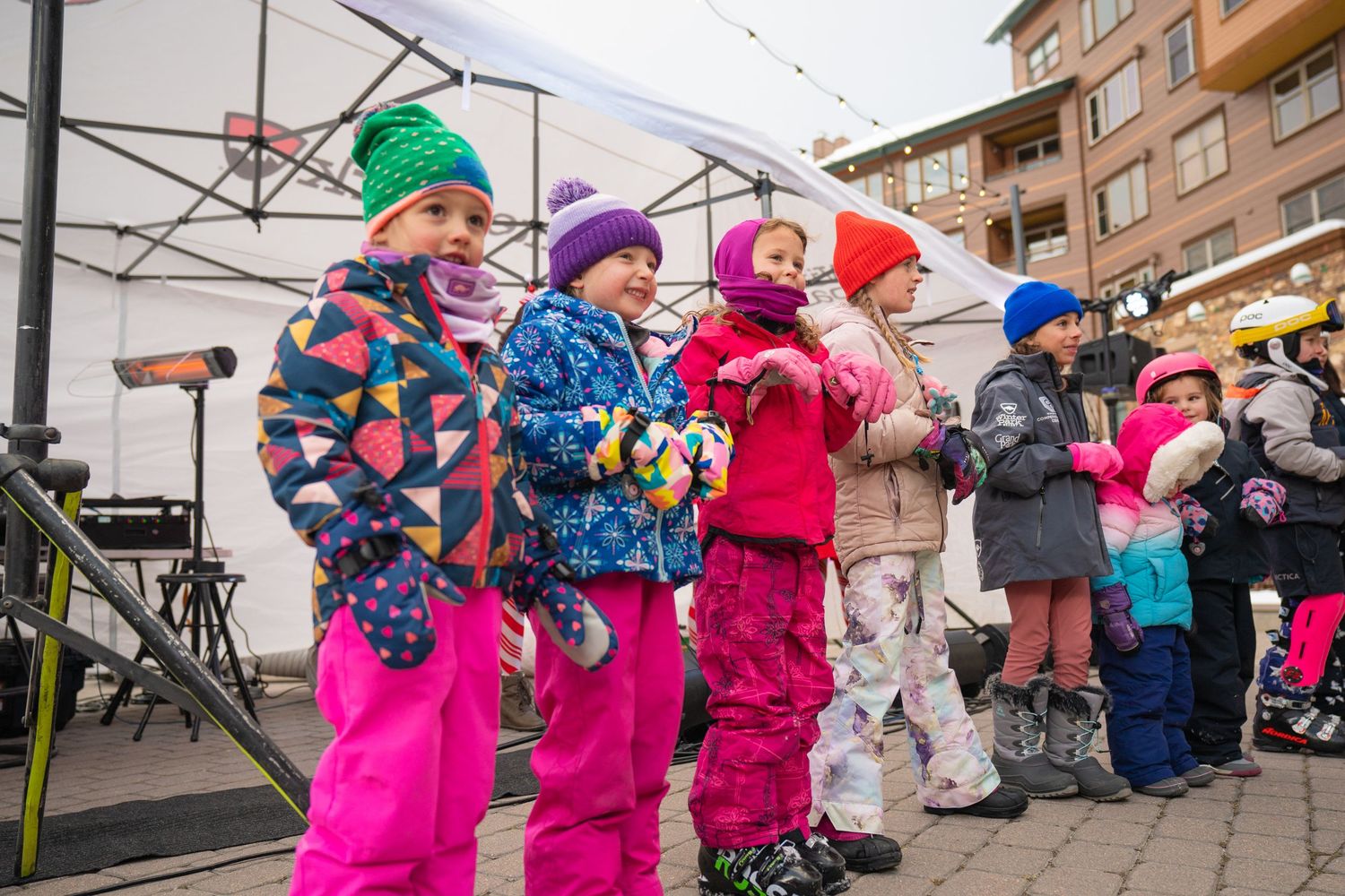 Children in colorful winter gear stand together at an outdoor winter event.