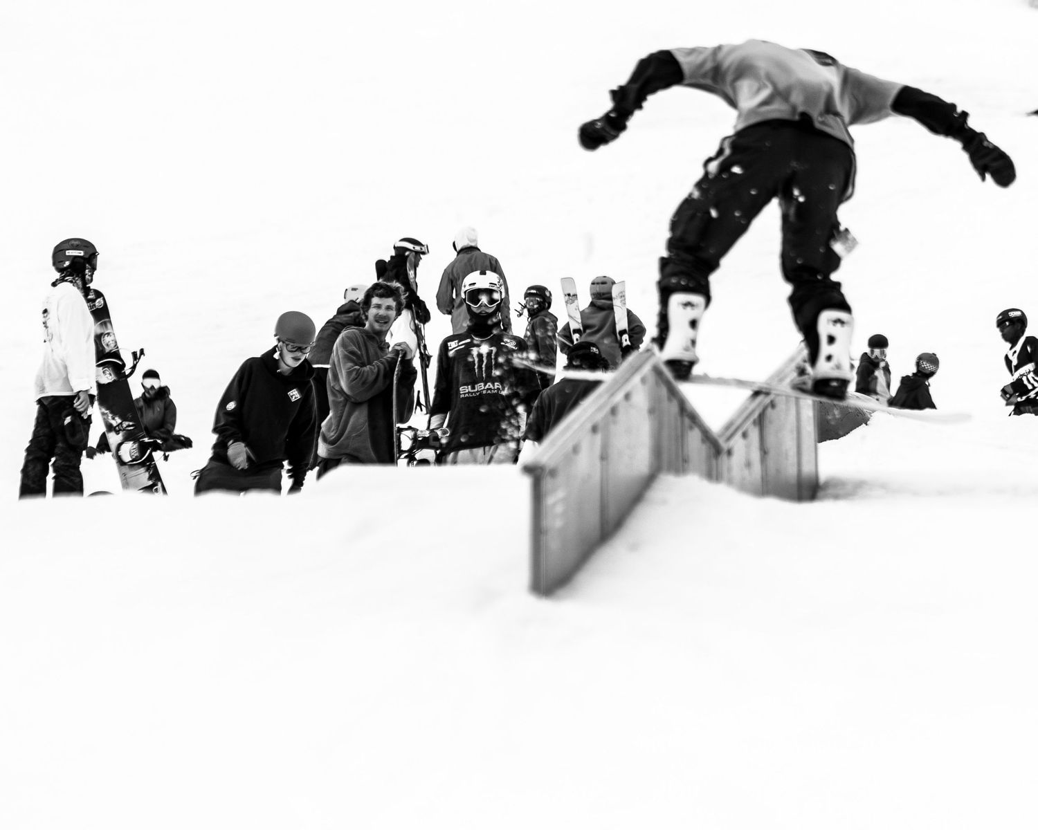 Black and white shot of a snowboarder performing an aerial trick over a rail with spectators in the background.