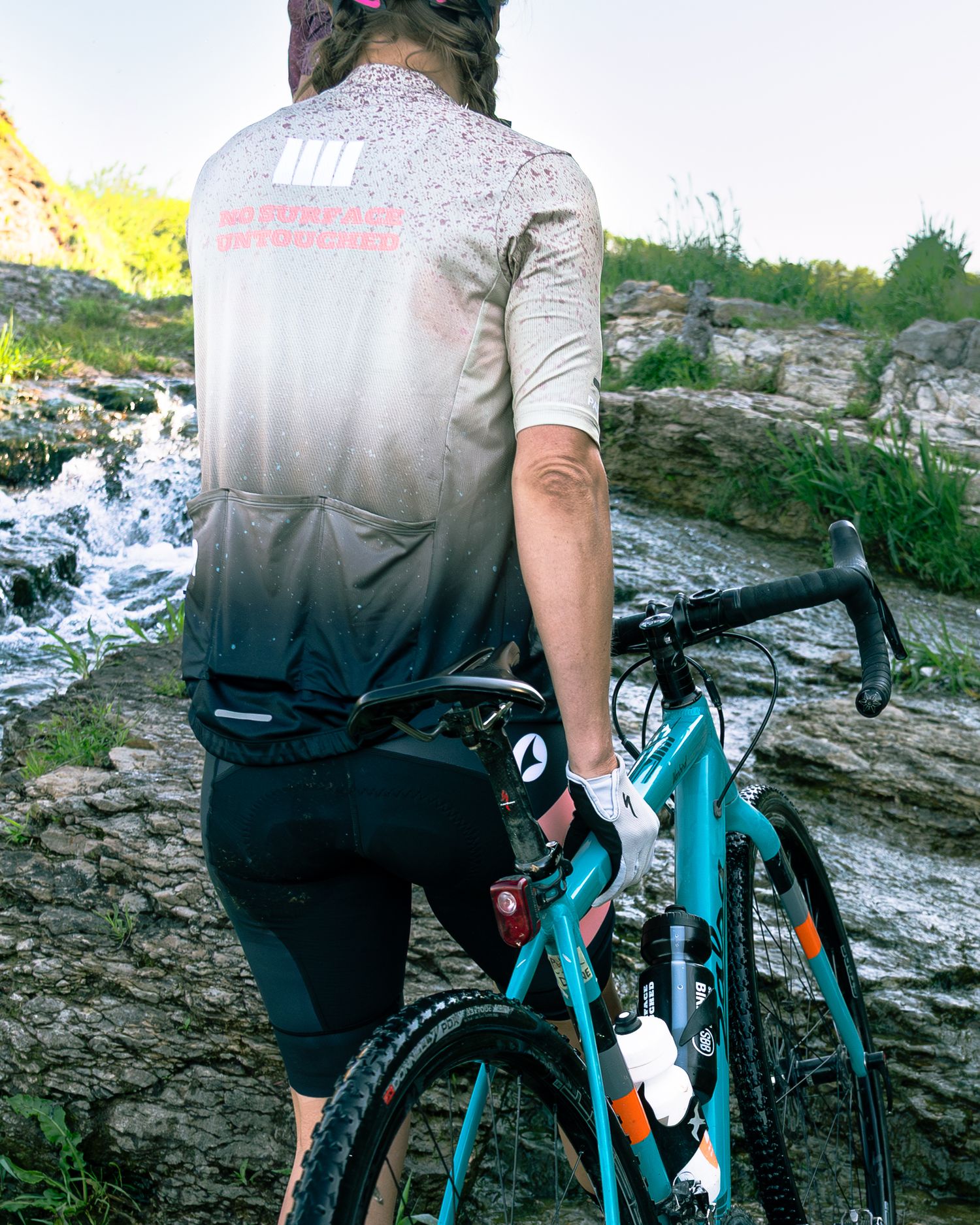 Cyclist in gradient jersey holds teal gravel bike near rocky waterfall on outdoor trail.