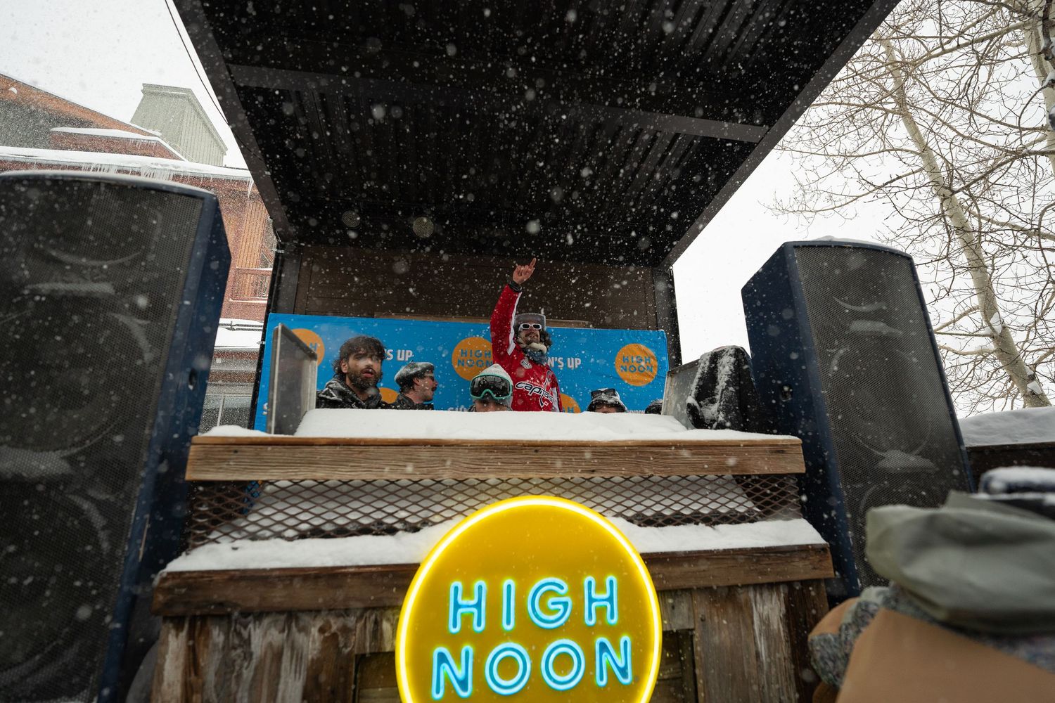 Snow falling on outdoor stage with High Noon sign and speakers during winter event.
