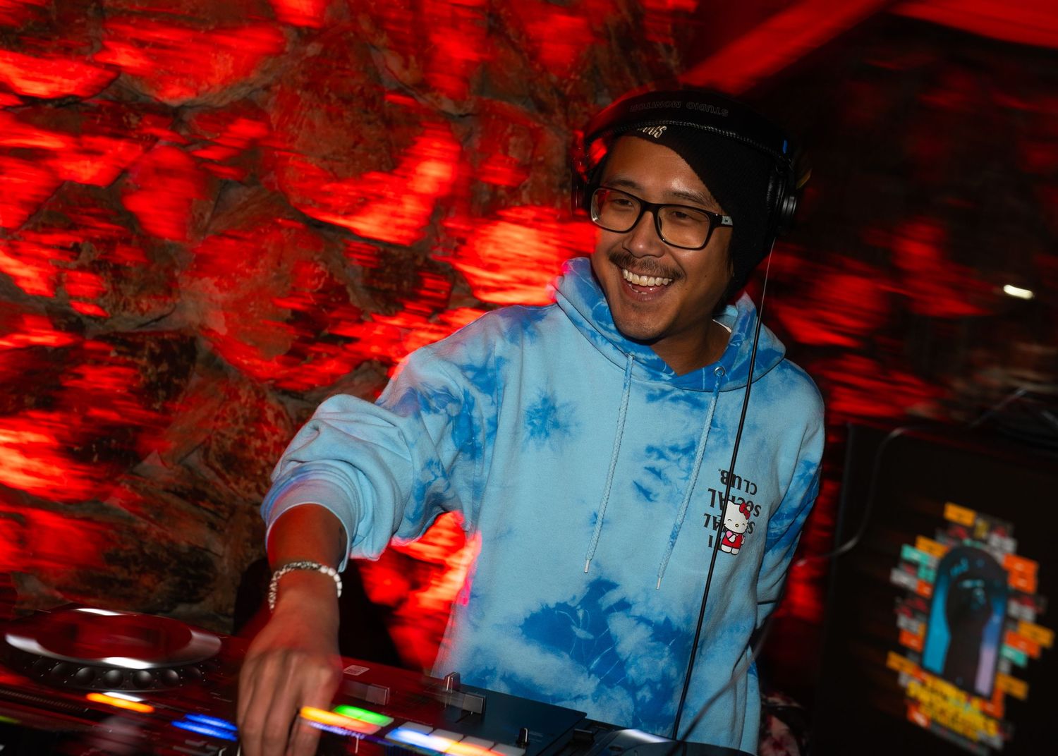 Smiling DJ wearing headphones and blue tie-dye hoodie mixing music at turntables under red ambient lighting.