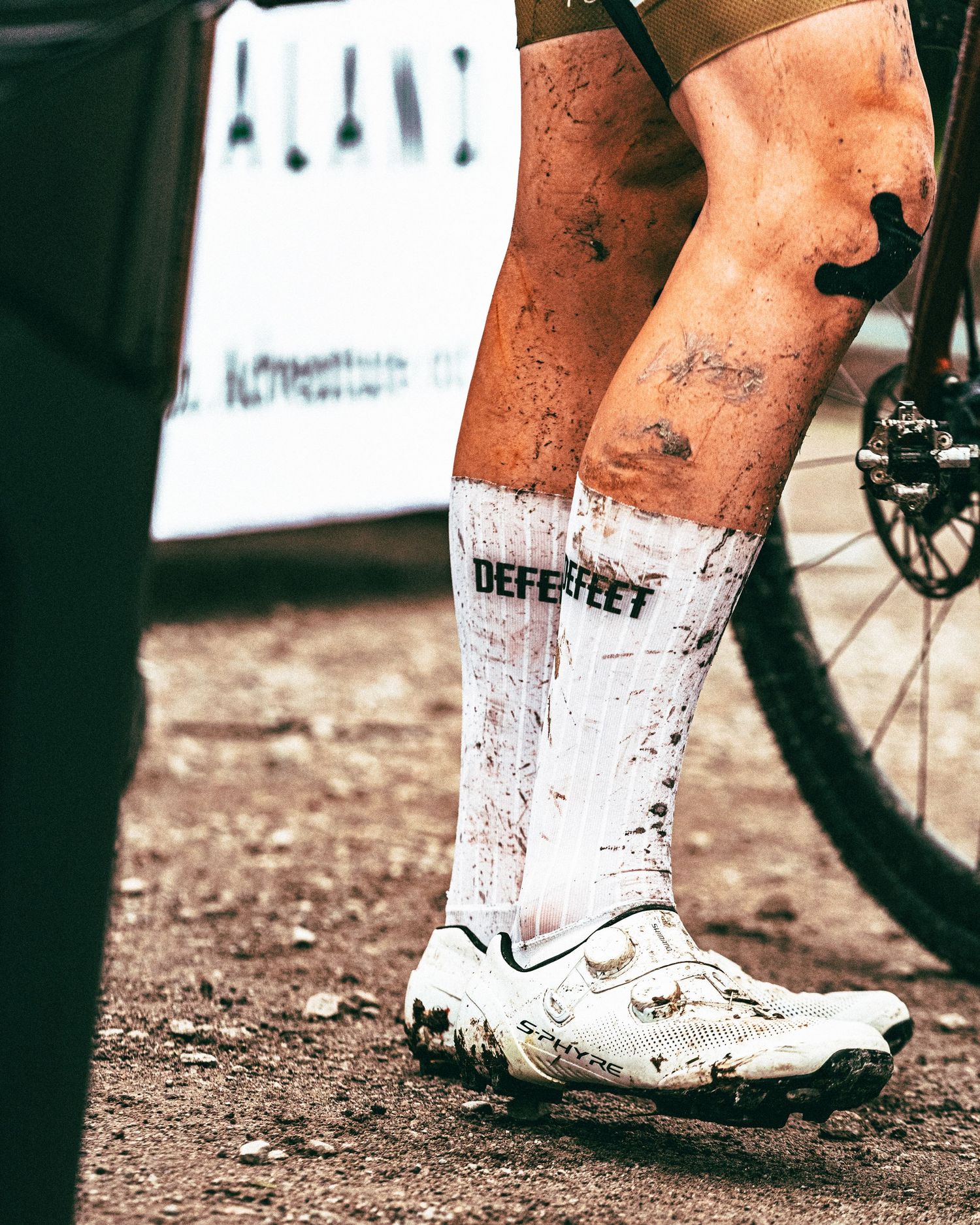 Muddy cyclist legs wearing white DEFEET socks and mountain bike shoes after an off-road race.