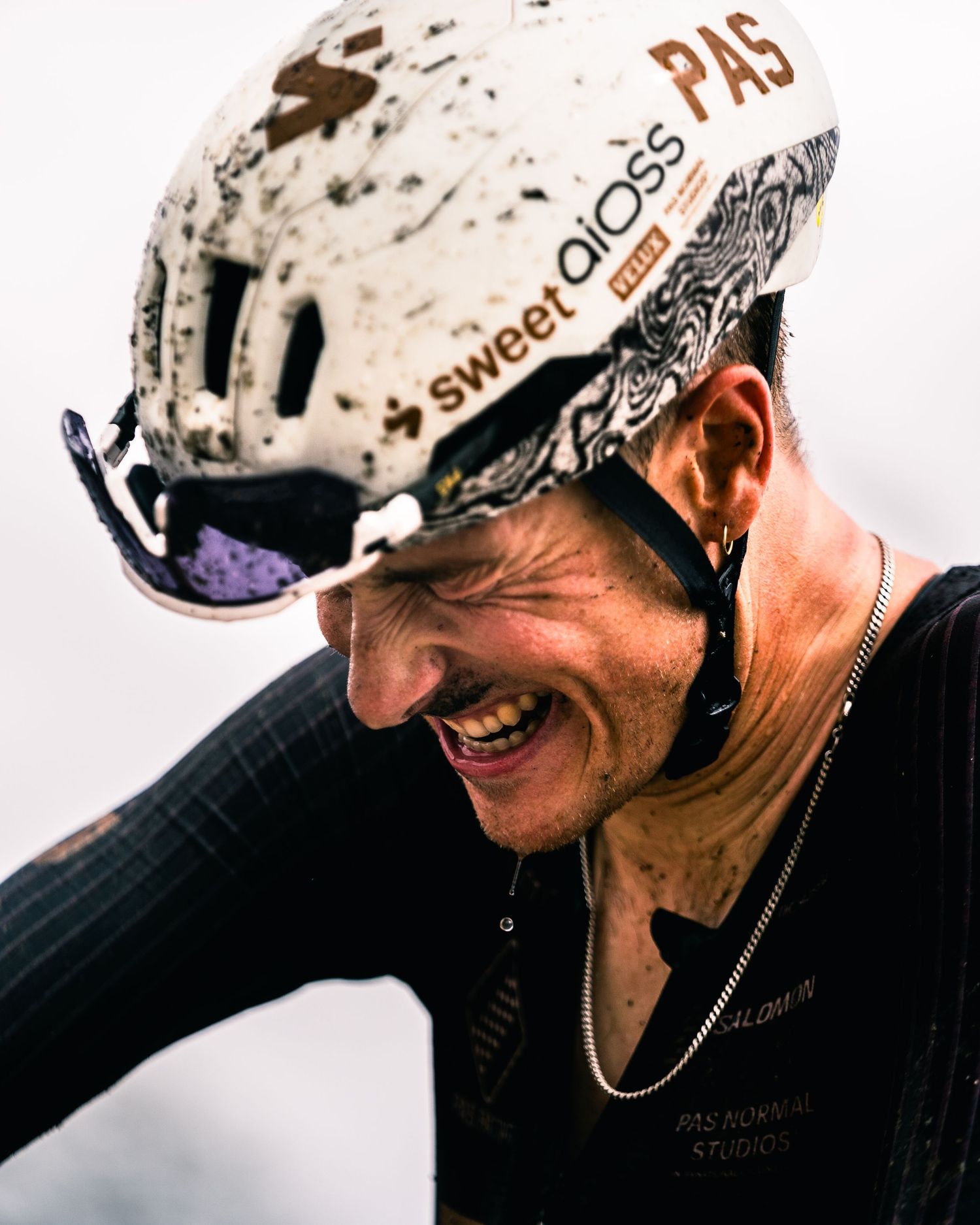 Smiling cyclist wearing a muddy white Sweet Protection helmet with PAS branding during an intense outdoor race.