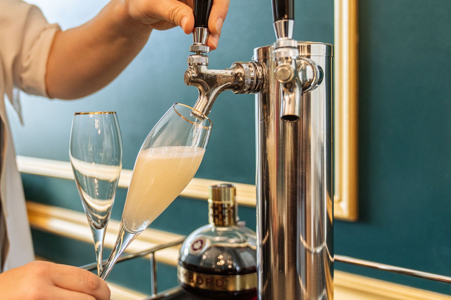 Draft beer is being poured from a stainless steel tap into a champagne flute at a bar with teal walls.