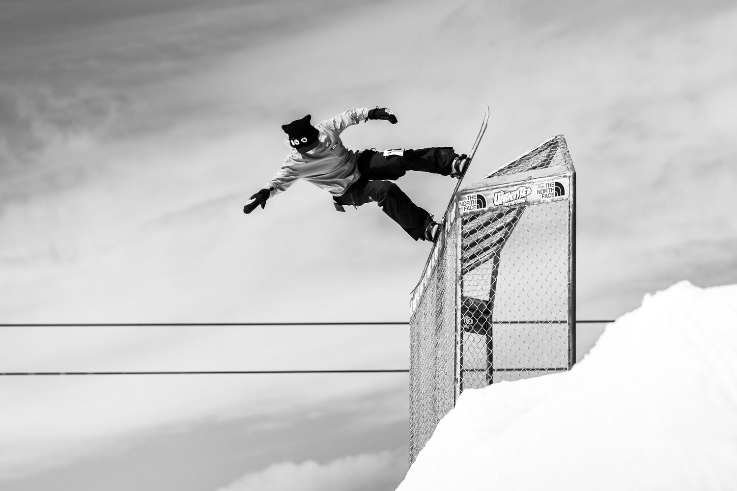 A snowboarder performs an aerial trick over a chain-link fence in a dramatic black and white action shot.