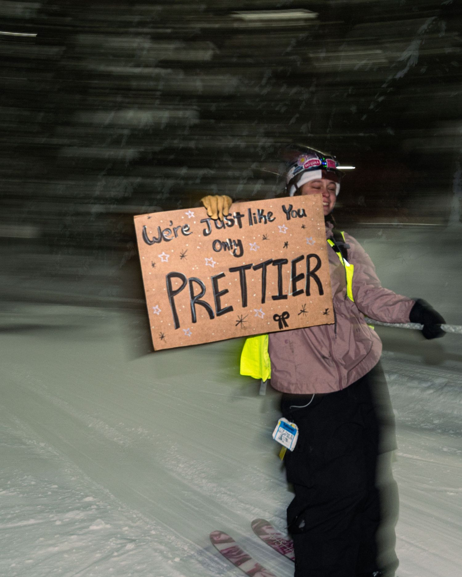 Snowboarder holding a cardboard sign reading We're Just Like You Only Prettier at a snowy night ski event.