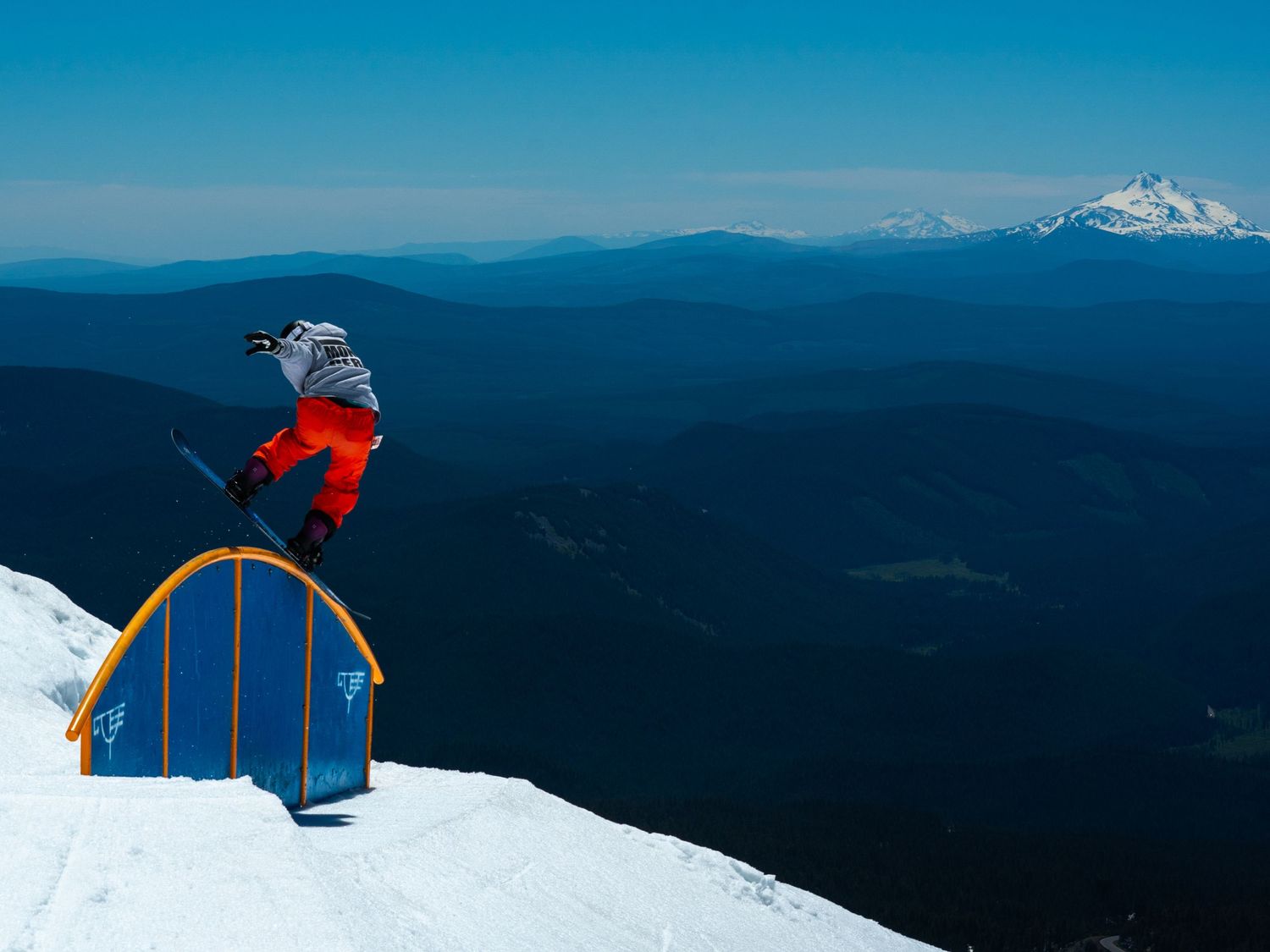Skier performs jump over terrain park rail with snow-capped mountains in background.