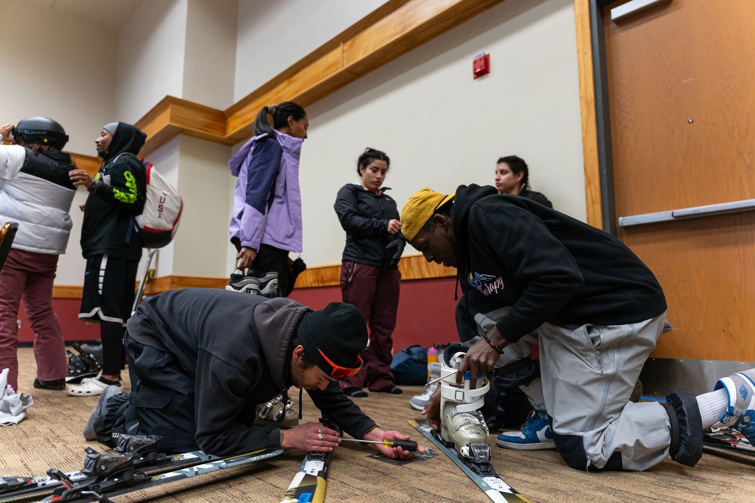 Skiers preparing equipment and gearing up in an indoor lodge space.