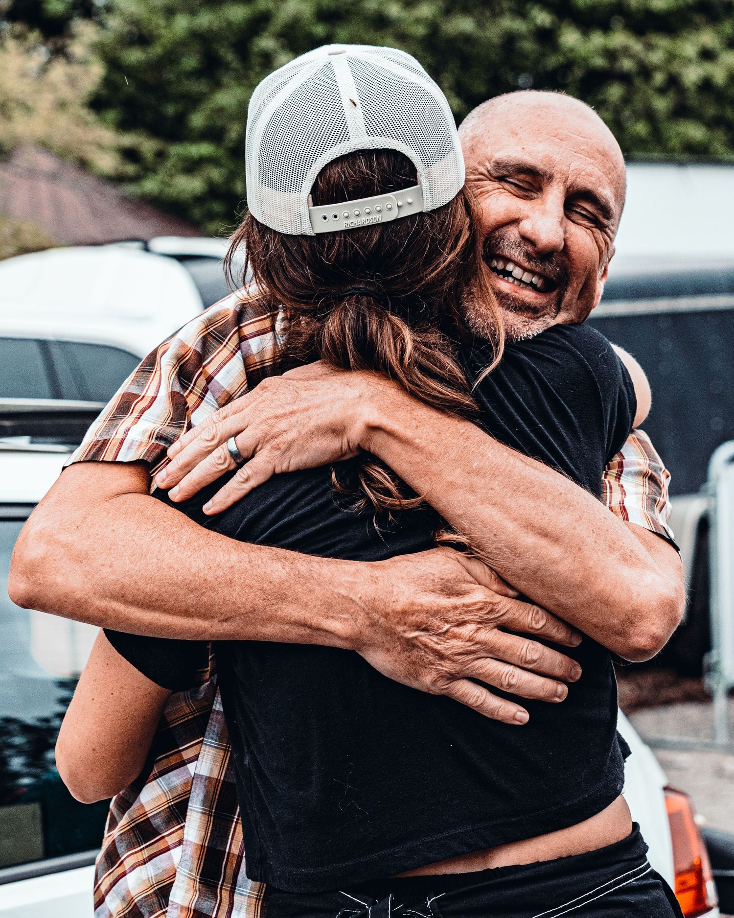 Two people sharing a warm hug outdoors, with a bald man smiling joyfully while embracing someone in a plaid shirt.