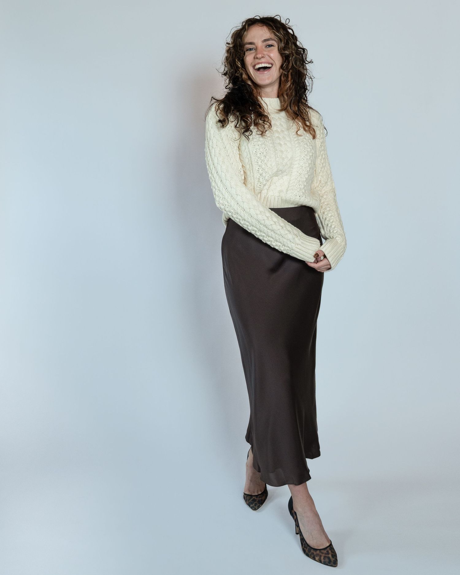 A person in a cream cable knit sweater and dark skirt poses playfully against a white studio backdrop.