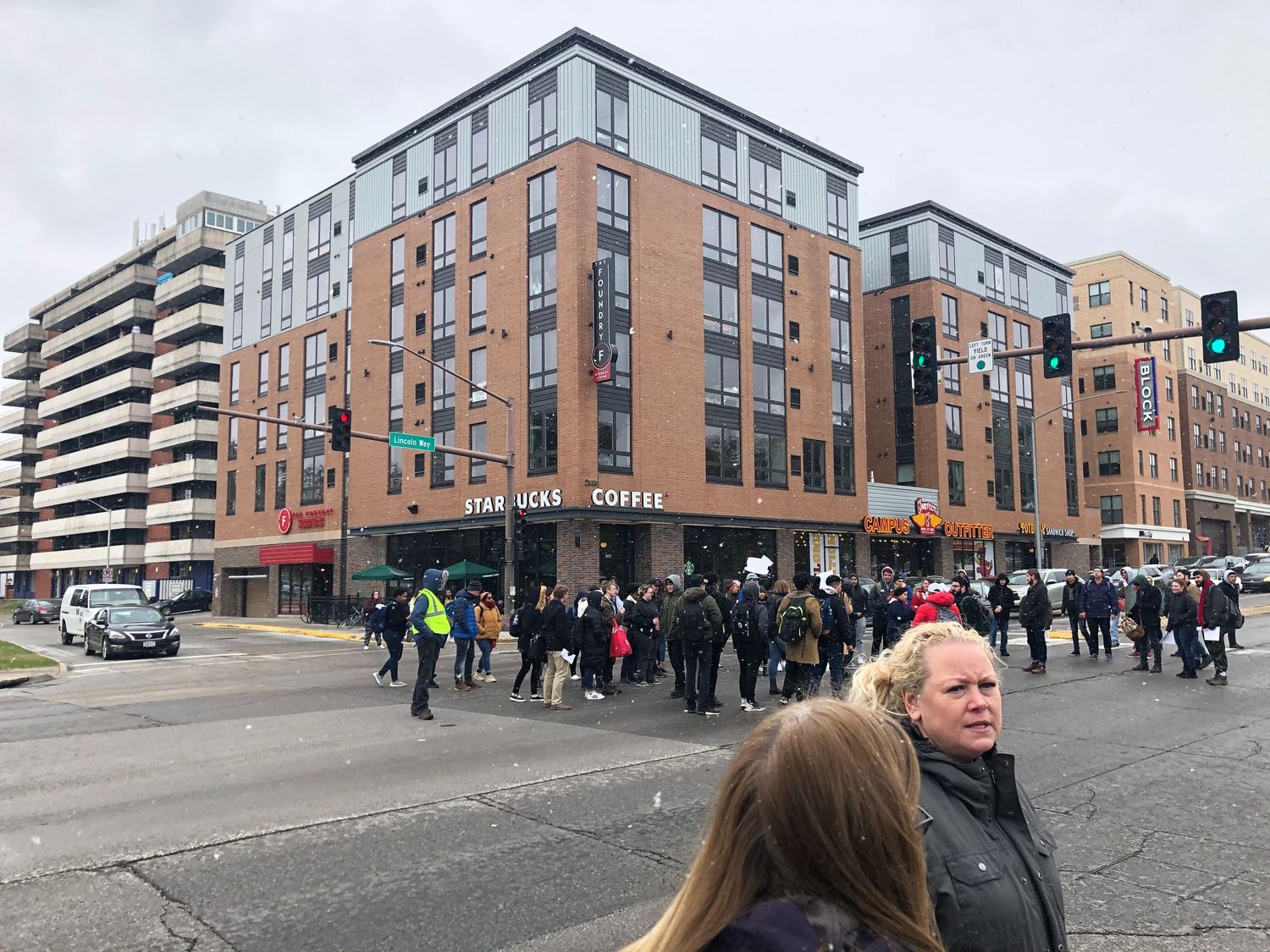 Pedestrians gather outside modern brick apartment buildings with retail space at street level in an urban setting.