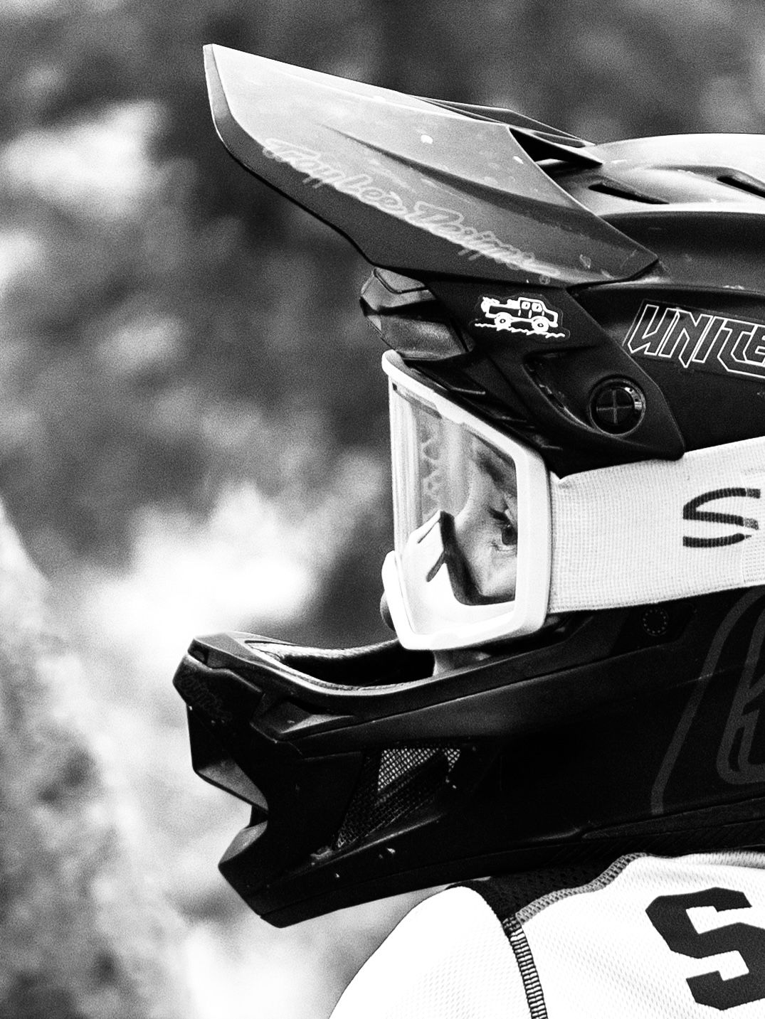 A close-up black and white photo of a motocross helmet and goggles against a blurred background.