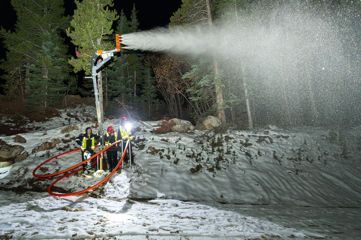 Snow-making equipment spraying artificial snow on dark mountain slope with crew monitoring operations.