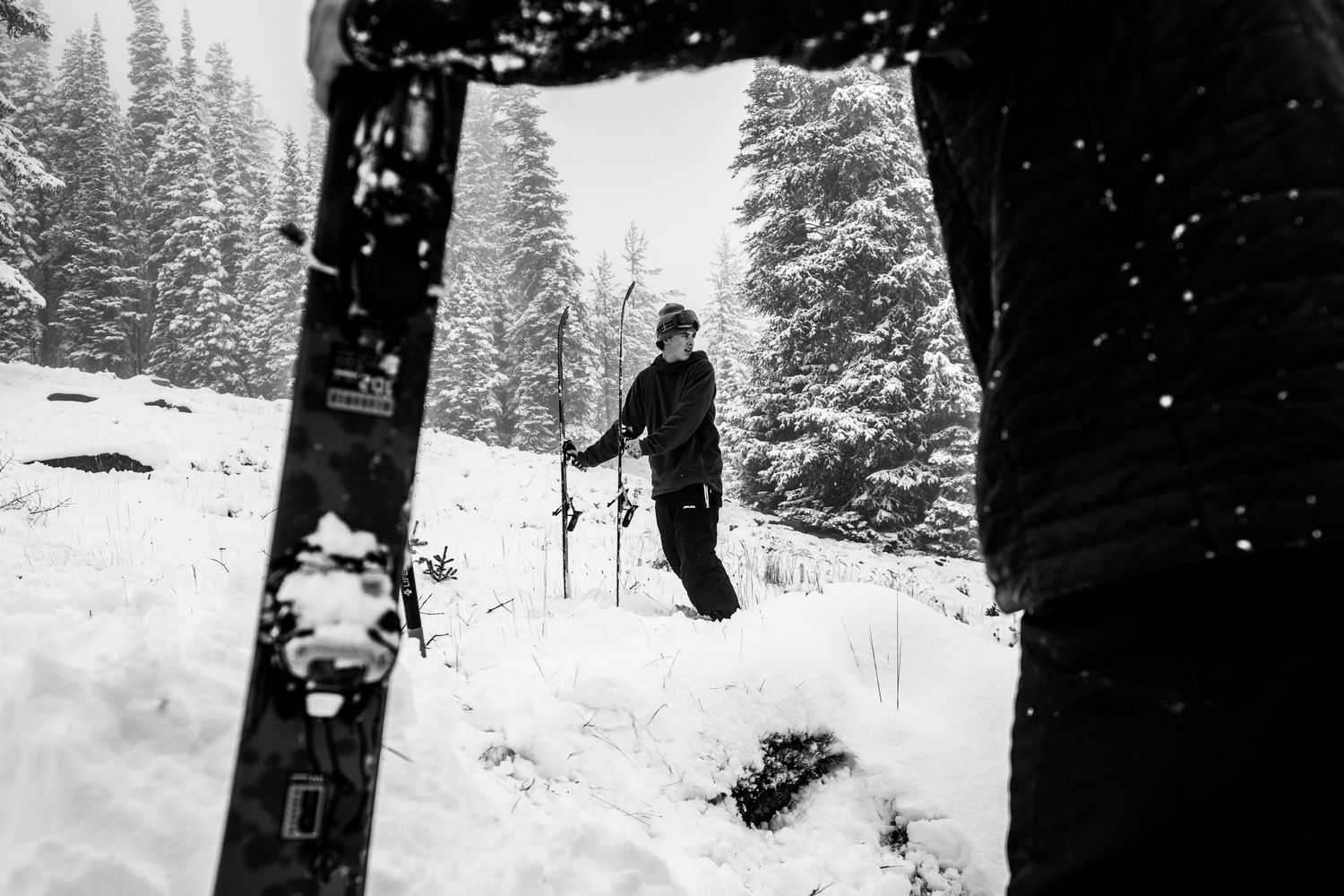 Black and white shot of skier hiking through snowy forest framed between trees.