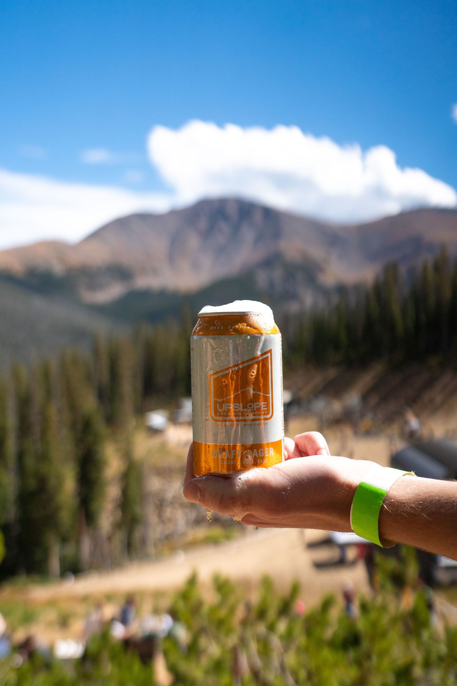 Hand holding a beer can against mountain landscape with blue sky and white clouds.
