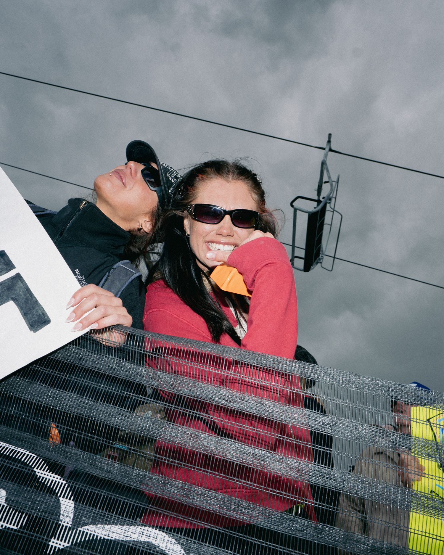 Two smiling spectators ride chairlift at snowy ski resort under dramatic cloudy skies.