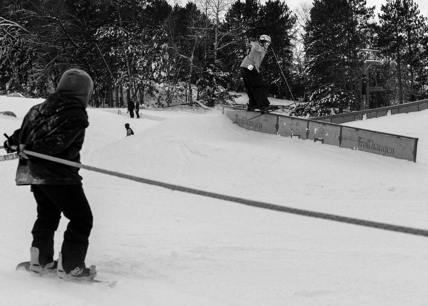 Black and white photo of young skier watching snowboarder perform rail trick at snow park.