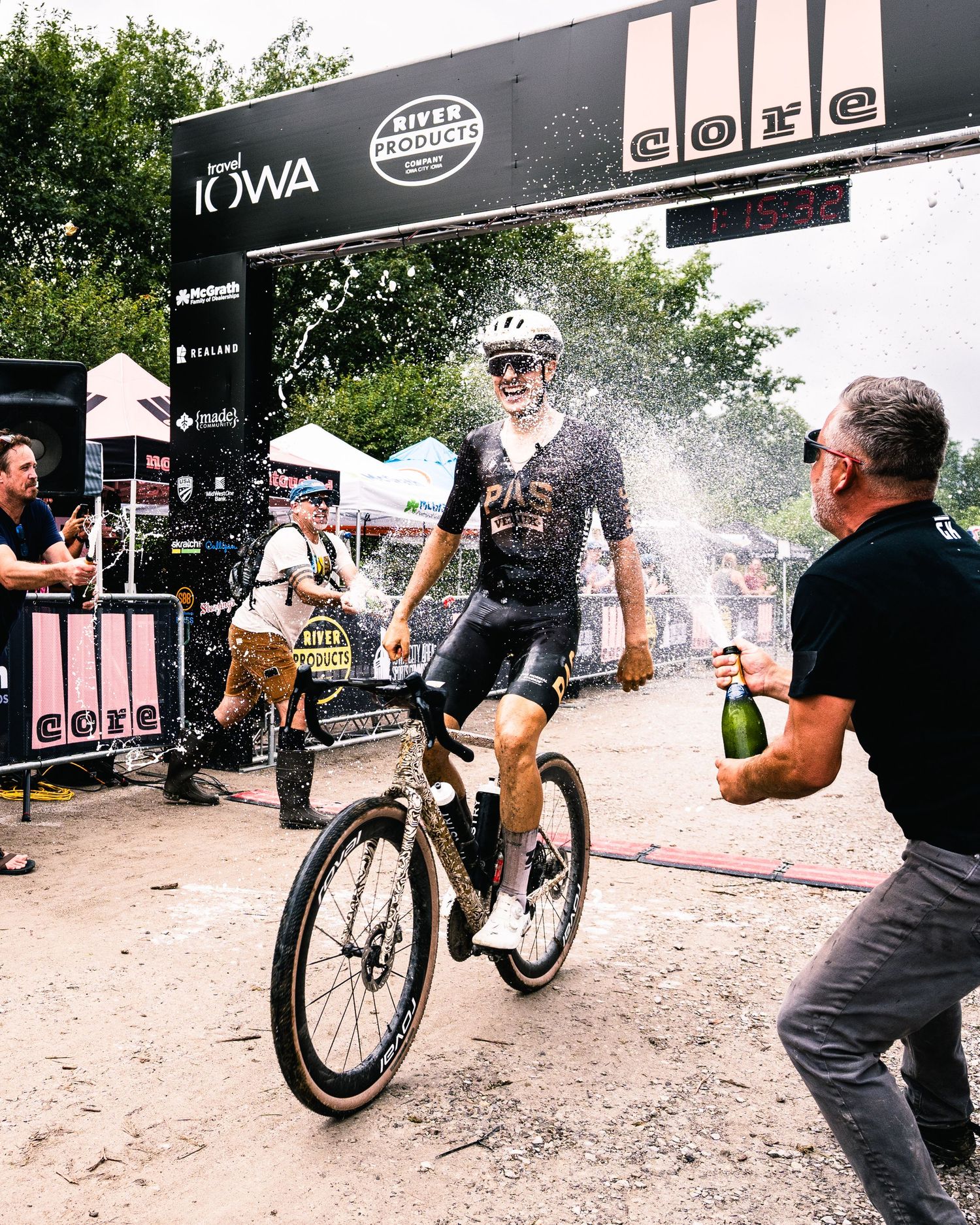 Cyclist crosses finish line at Iowa gravel race while spectator sprays champagne in celebration.