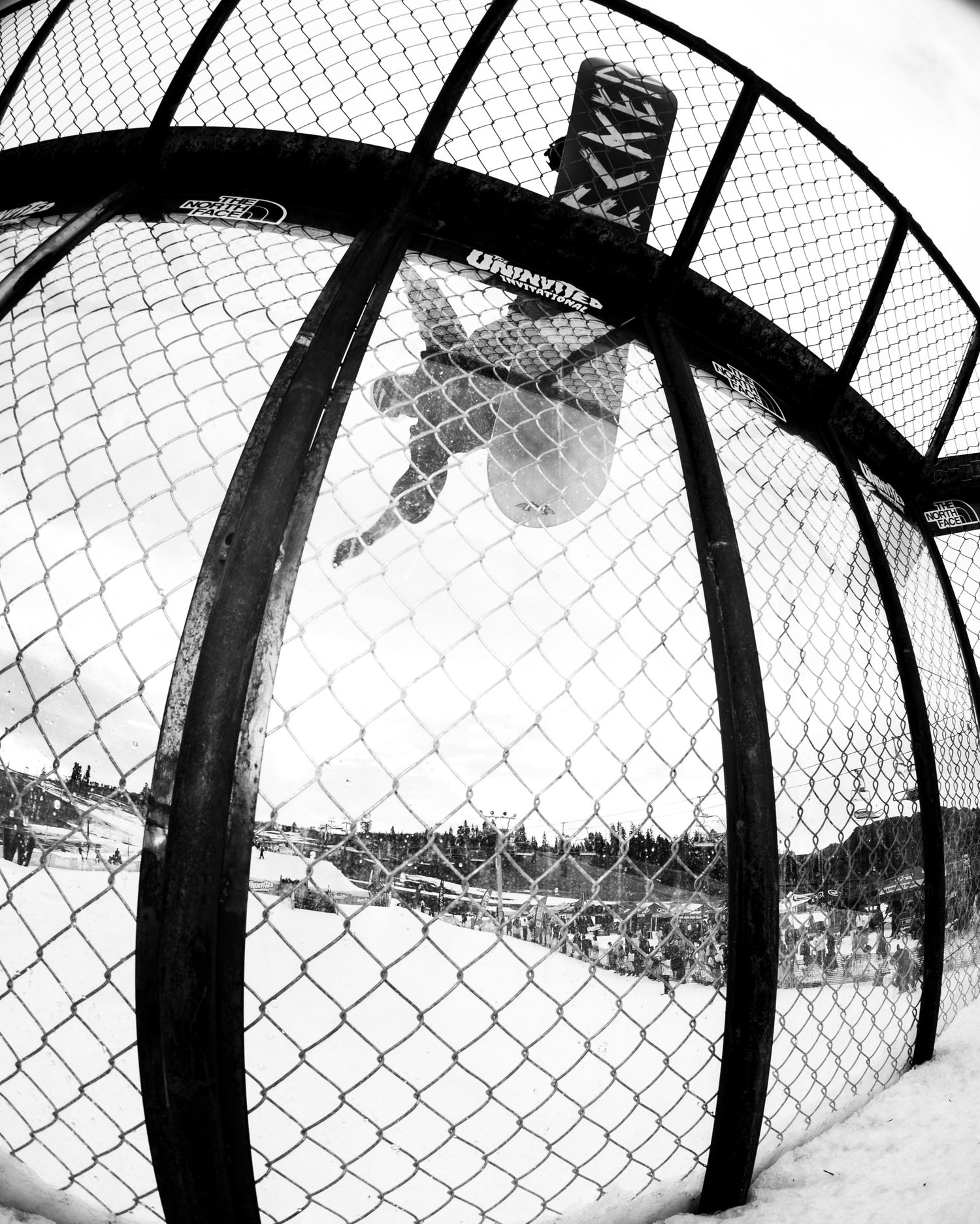 Black and white fisheye photo of a snowboarder performing an aerial trick inside a chain-link fence halfpipe.