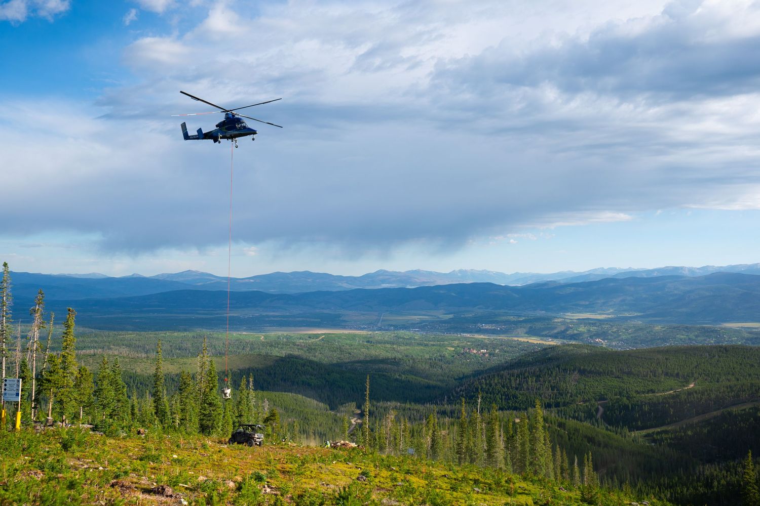 Military helicopter flies over mountainous landscape covered in dense forest under cloudy sky.