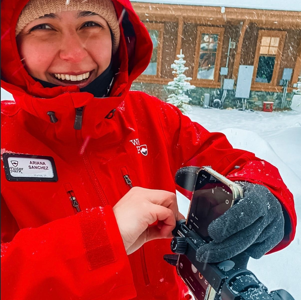 Winter outdoor sports enthusiast in bright red jacket at snowy ski resort adjusts equipment in front of rustic building.