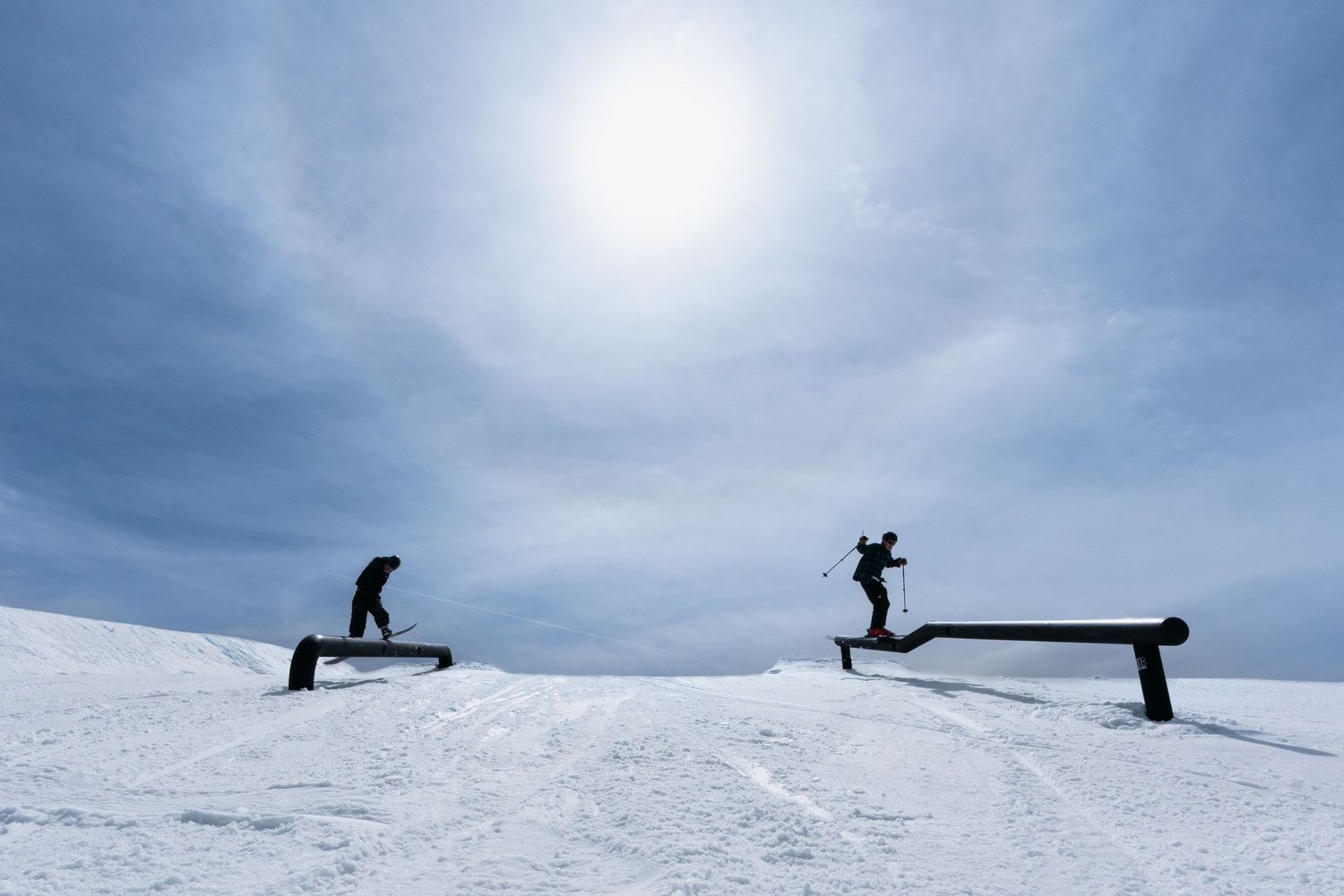 Two snowboarders performing tricks on rails in a snowy ski park under a bright sun and cloudy sky.