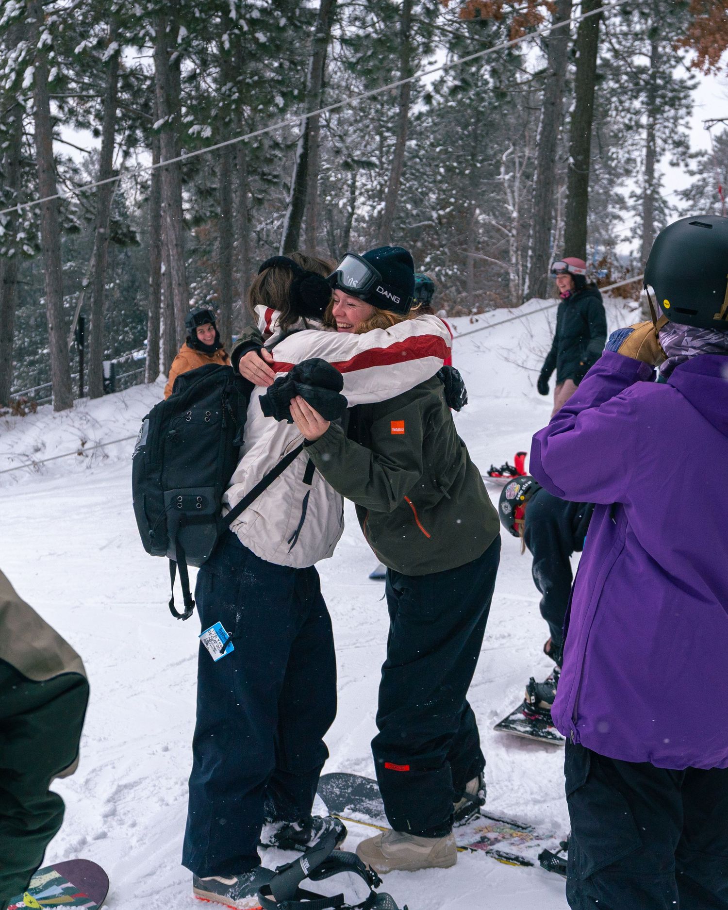 Two snowboarders share a warm hug on a snowy ski slope surrounded by snow-covered trees and friends.