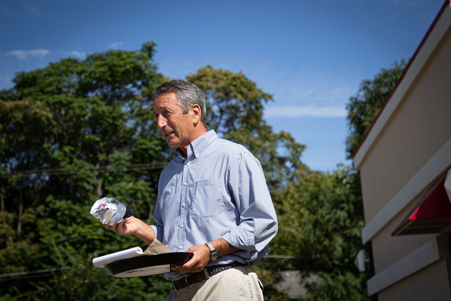 Man in blue button-down shirt holds sandwich while standing outdoors on a sunny day.