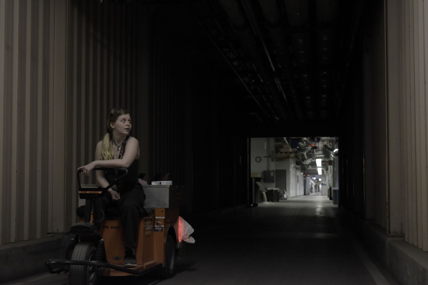Person sitting alone in dark warehouse corridor with distant light visible through doorway.
