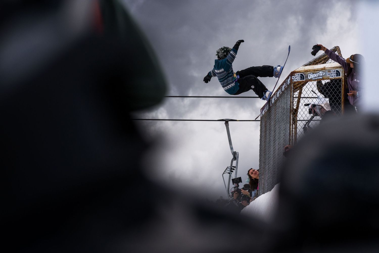 Snowboarder performs dramatic aerial trick over metal rail against cloudy sky.
