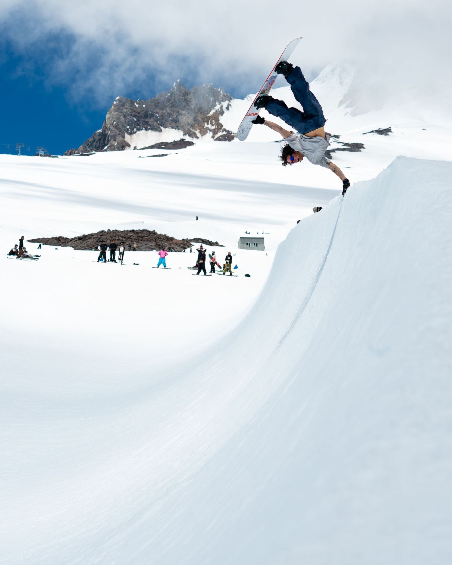 Snowboarder performing inverted aerial trick off large snow jump against blue sky.