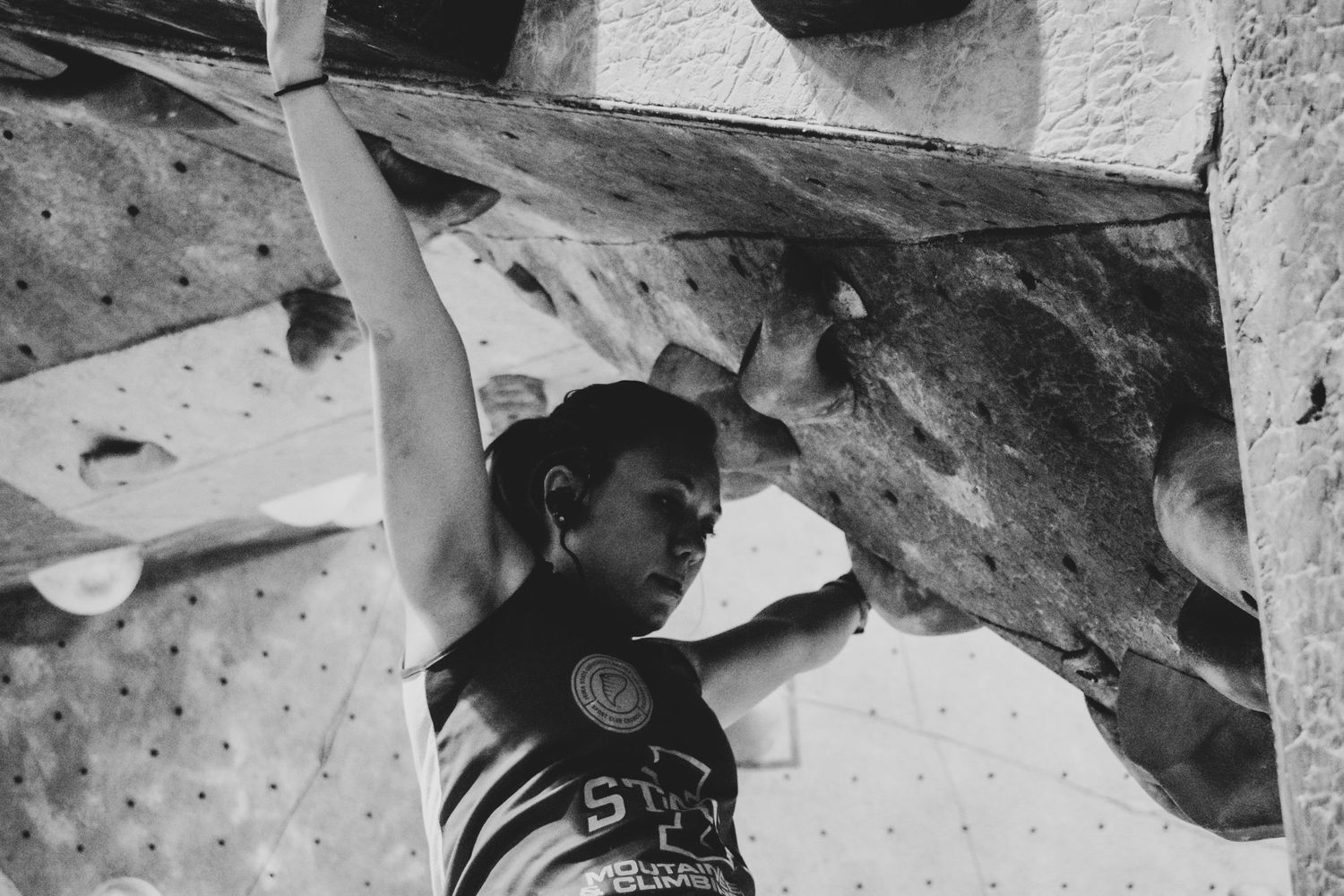 Indoor climber stretching upward on an angled climbing wall in dramatic black and white.