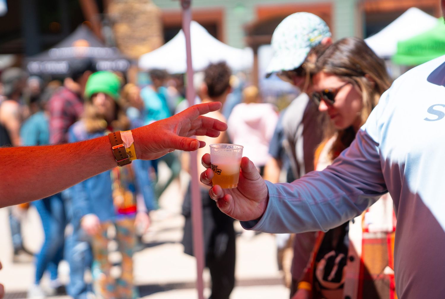 Hands exchanging a beverage sample at a crowded outdoor festival.