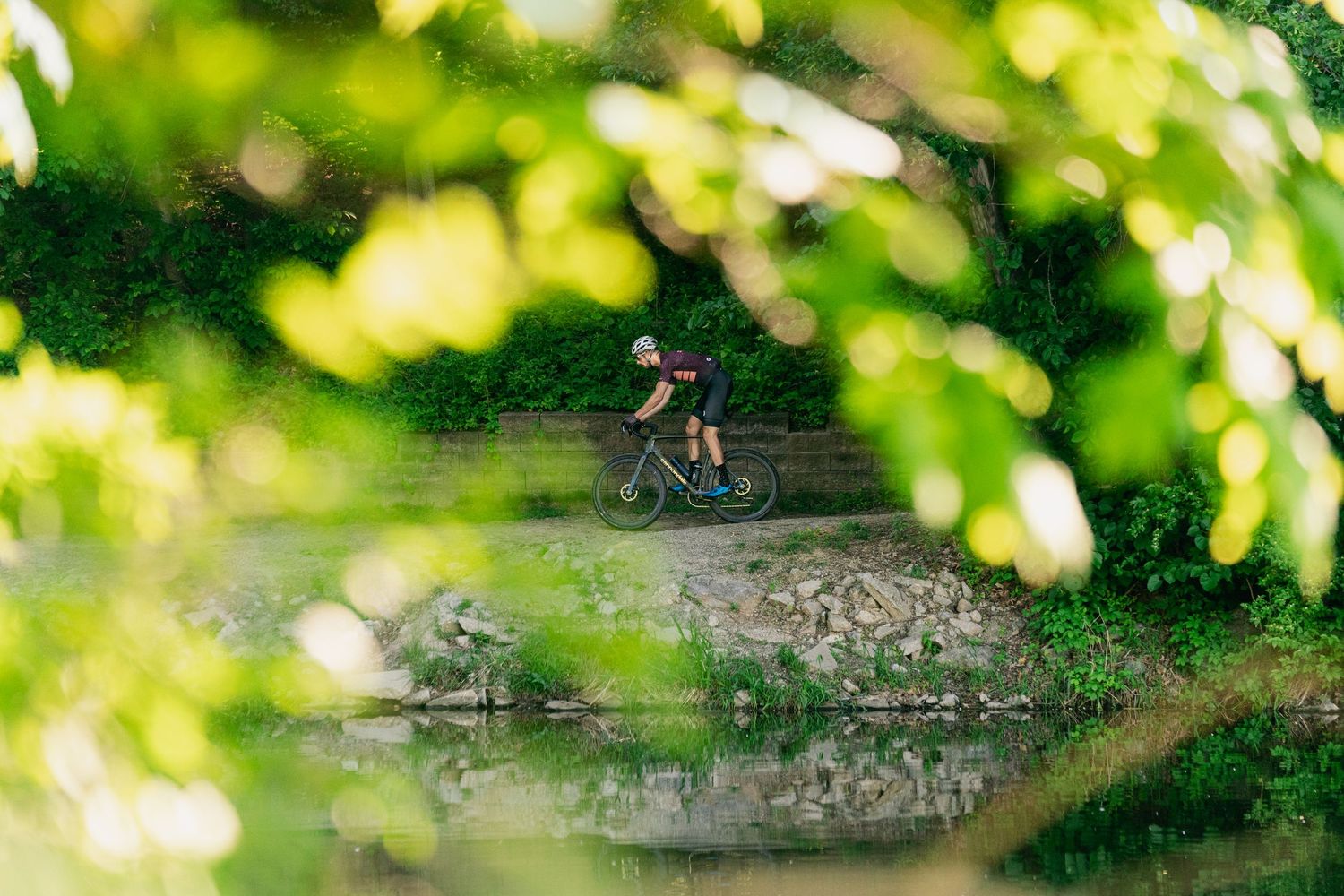 Cyclist riding a bike along a scenic green waterway path surrounded by lush foliage and trees.