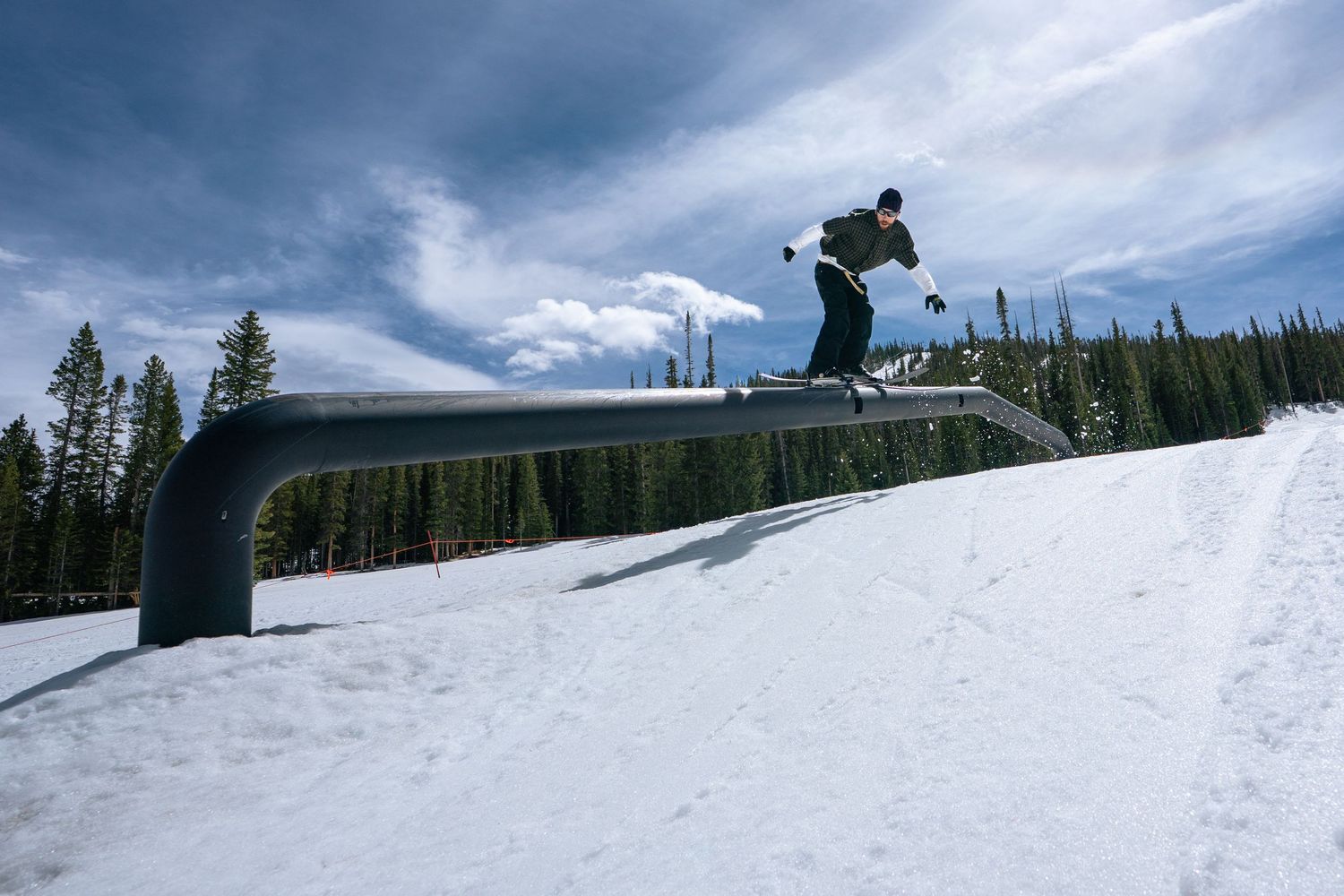 Snowboarder performing a rail grind trick on a metal rail at a ski resort terrain park with snowy slopes.