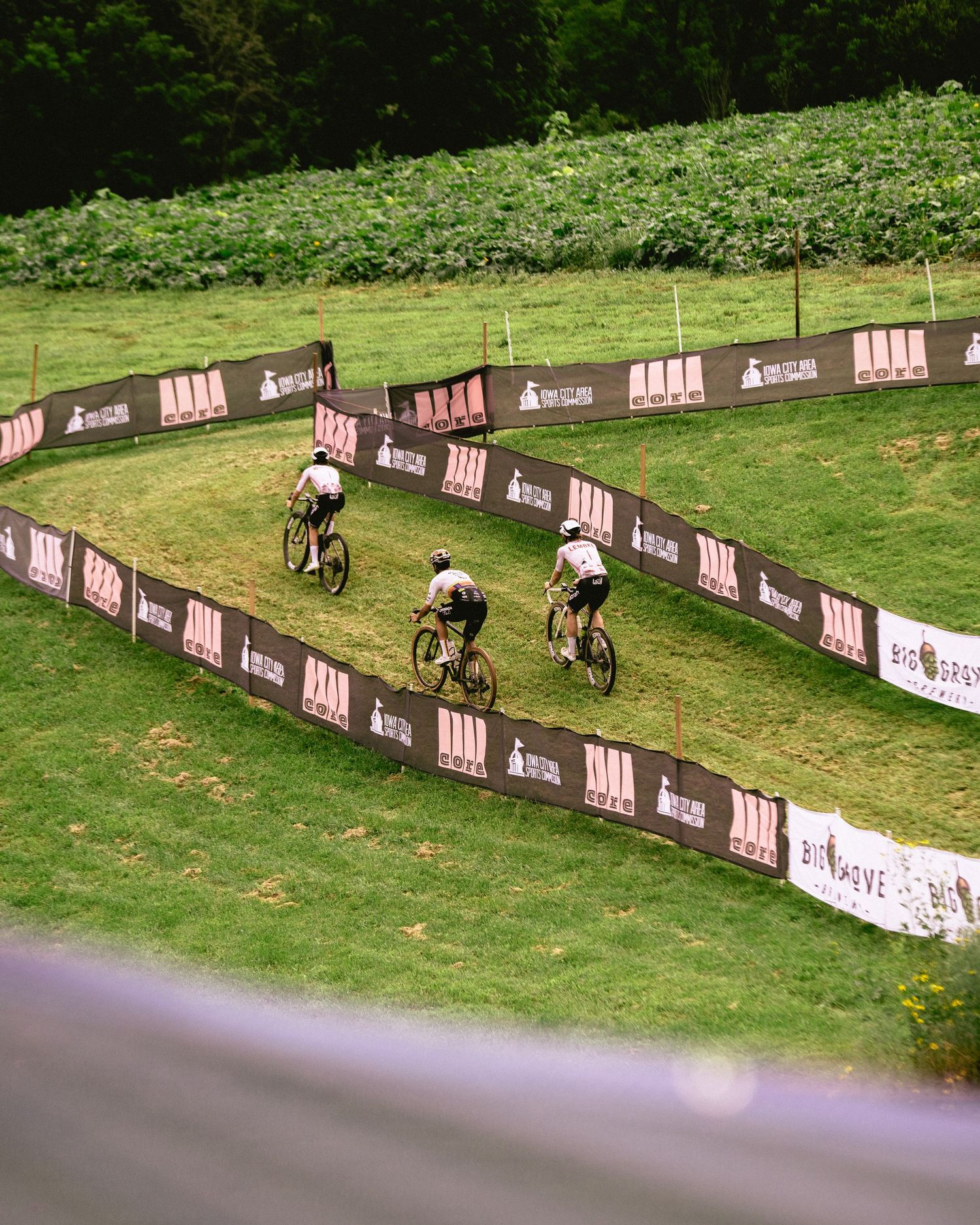 Three cyclists race on a winding grass course lined with sponsor banners at an outdoor mountain bike event.