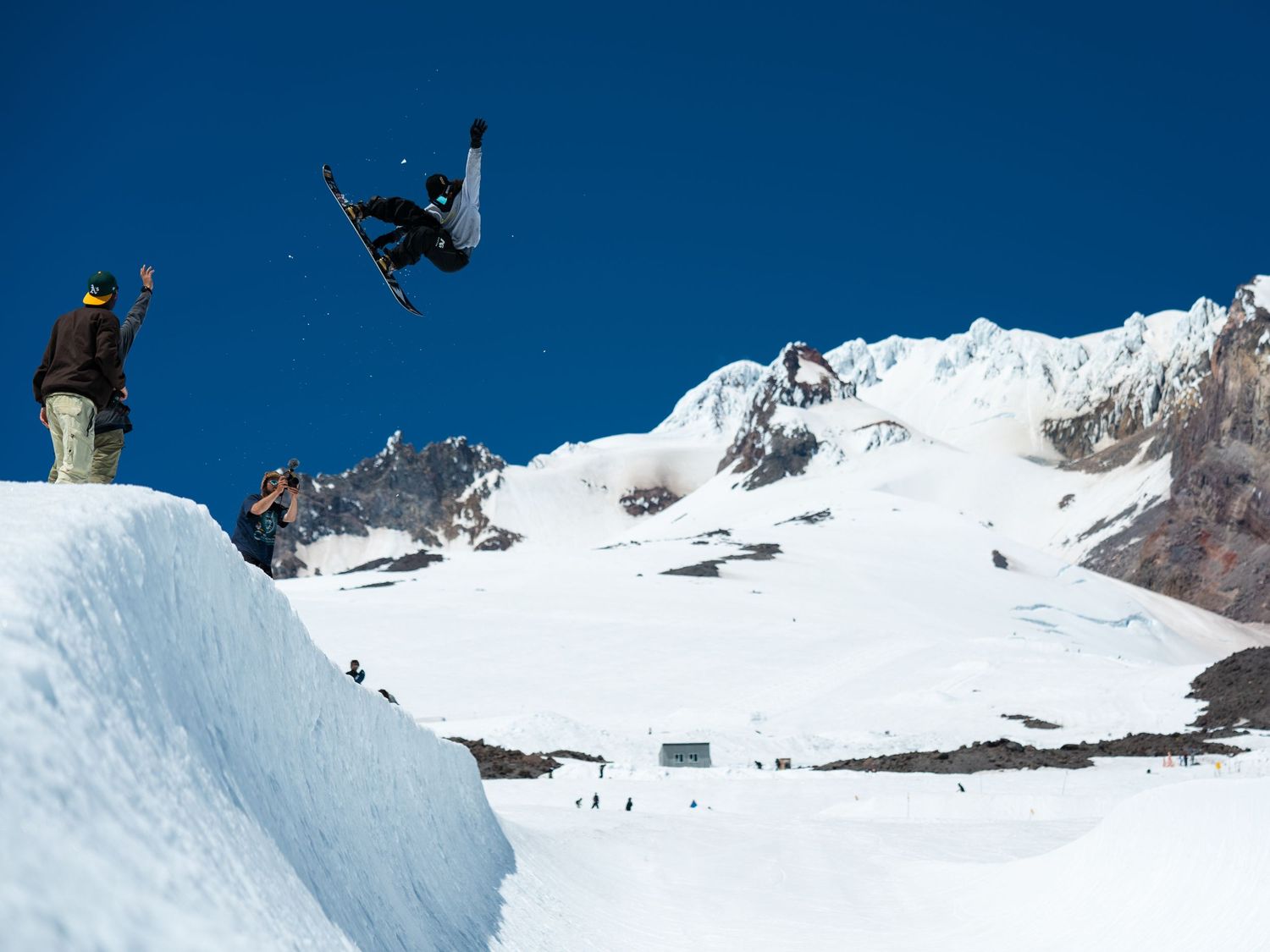 Snowboarder performing a dramatic aerial jump against blue sky and snowy mountain backdrop.