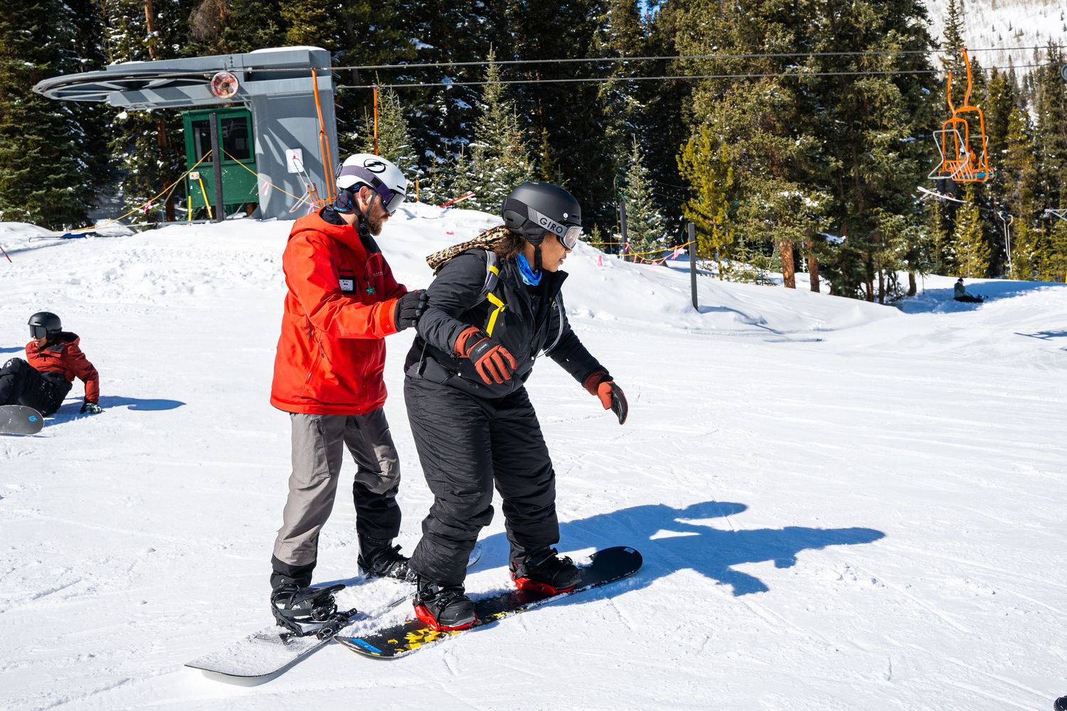 Snowboarding instructor assisting beginners on gentle snowy slope with ski lifts visible.