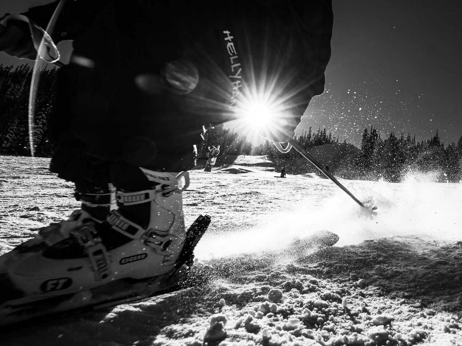 Black and white close-up of ski boots in motion spraying snow with sunburst in background.